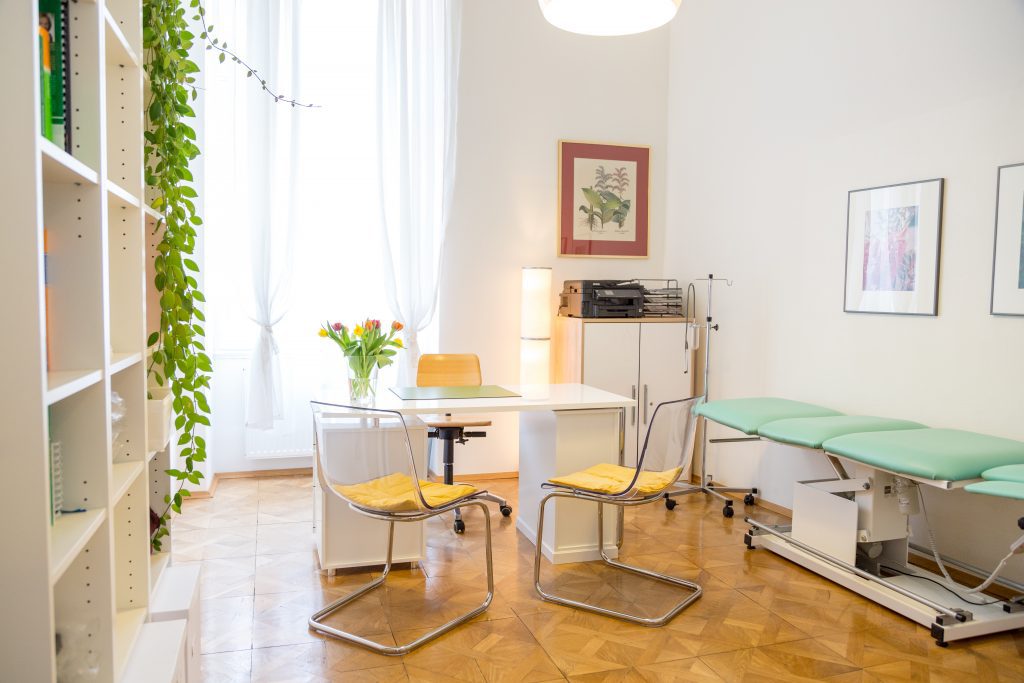 Das Foto zeigt einen der Therapieräume: ein heller, freundlicher Raum mit einem Tisch und zwei Sesseln in der Mitte, einem Regal auf der Linken und einer Therpieliege auf der rechten Seite des Raumes.